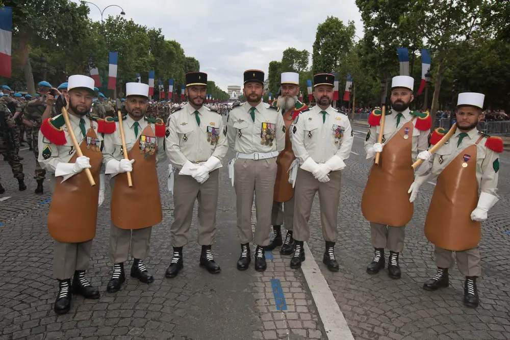 Pionniers de la Légion étrangère en tenue traditionnelle avec barbe, tablier de cuir et hache à l'épaule lors d'un défilé.