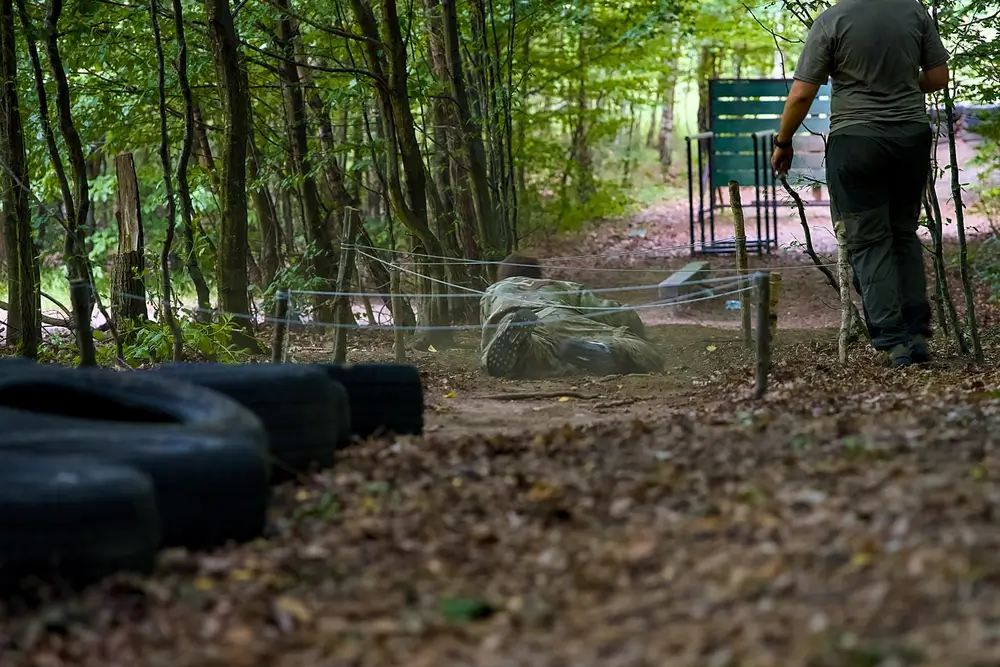Candidat en treillis rampant sous un obstacle lors d'un test sportif militaire de sélection en forêt.