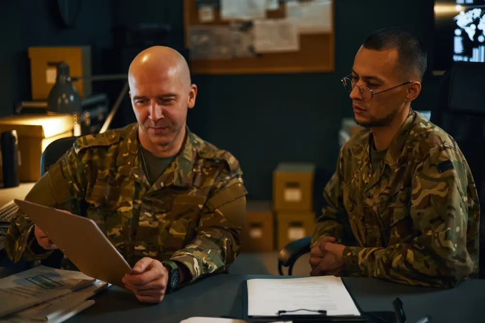 Deux sous-officiers militaires en uniforme analysent un document lors d'un briefing dans une salle de commandement.
