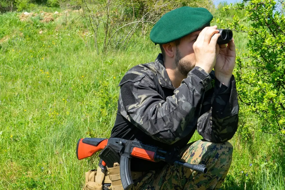 Soldat au béret vert orné de l'insigne, observant aux jumelles en plein champ lors d'un exercice militaire.