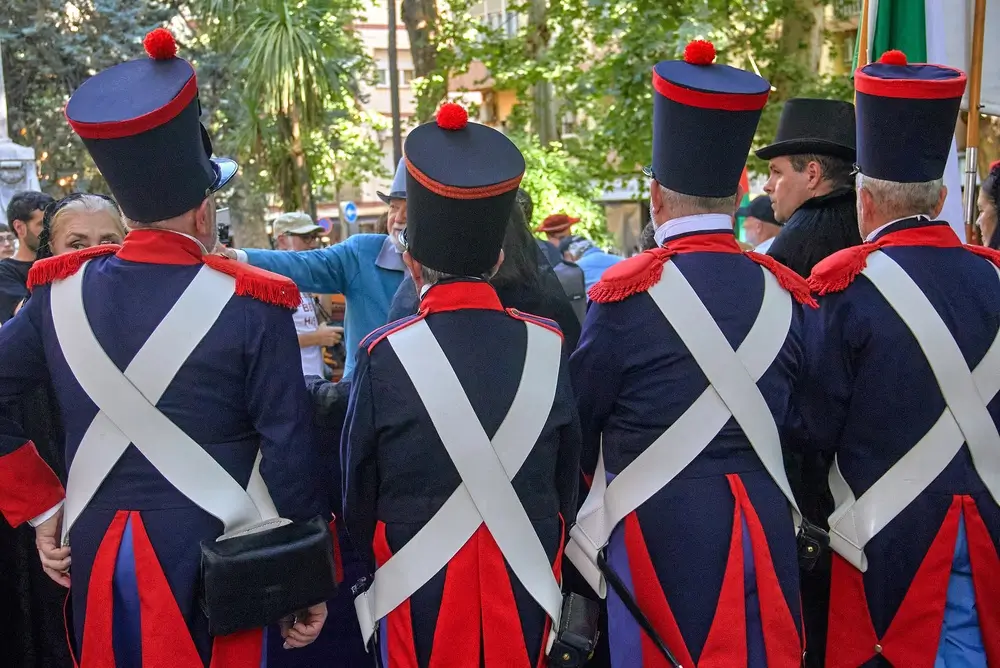 Soldats de dos en uniforme militaire de cérémonie avec épaulettes rouges, baudriers blancs et ceinture bleue.